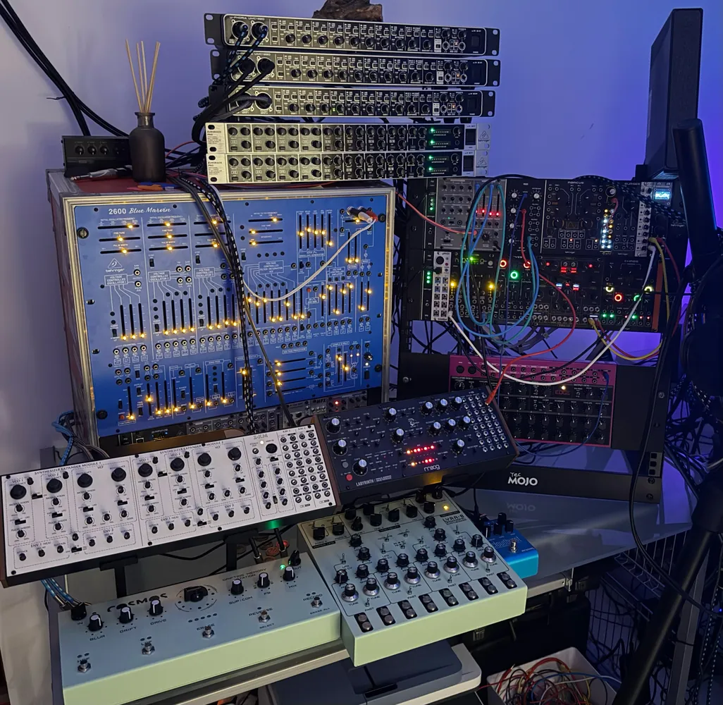 A rack of synthesiser modules lit in blue, with cream-coloured synthesisers on the desk below.