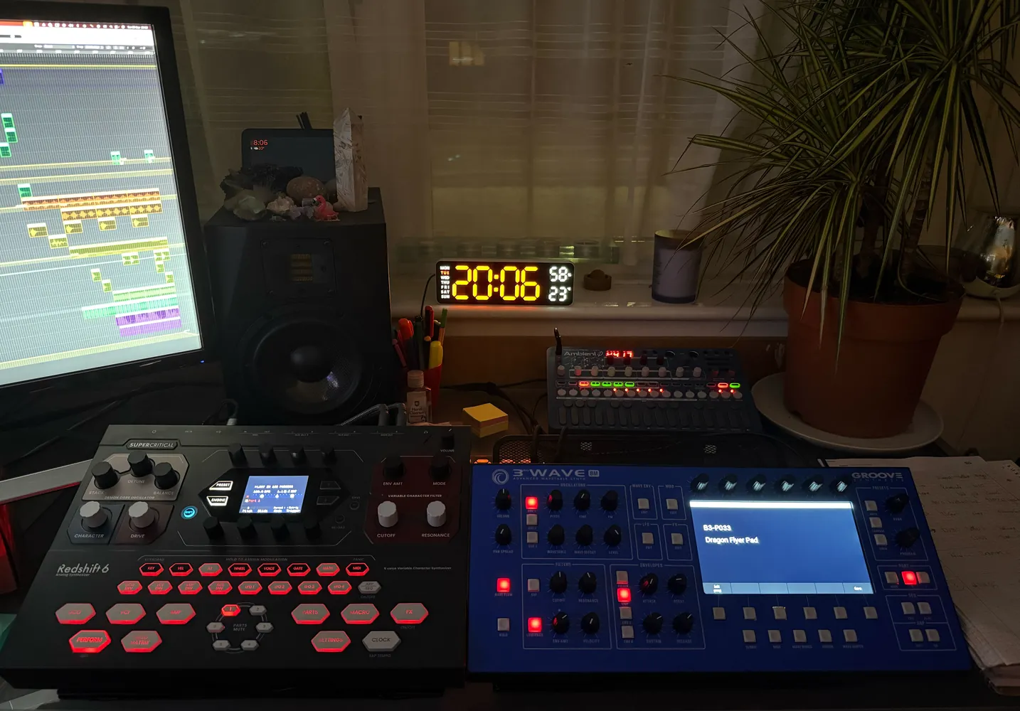 A drum machine and a blue synthesiser on the desk, with a monitor, a speaker, a clock and a plant behind them.