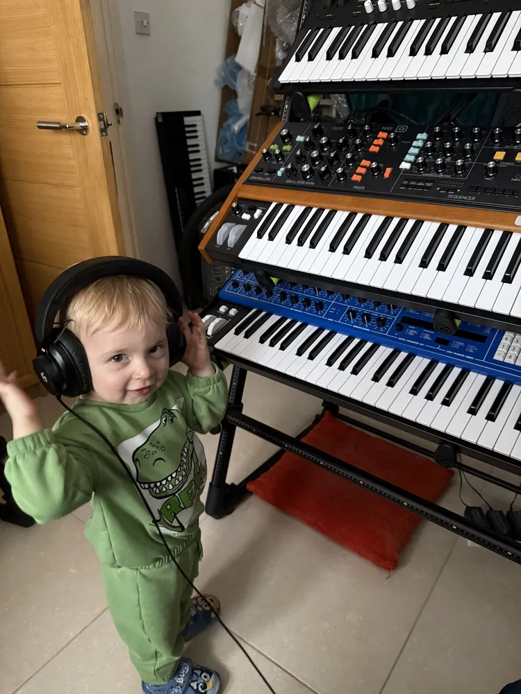 A small boy in big headphones dancing beside the stack of keyboards.